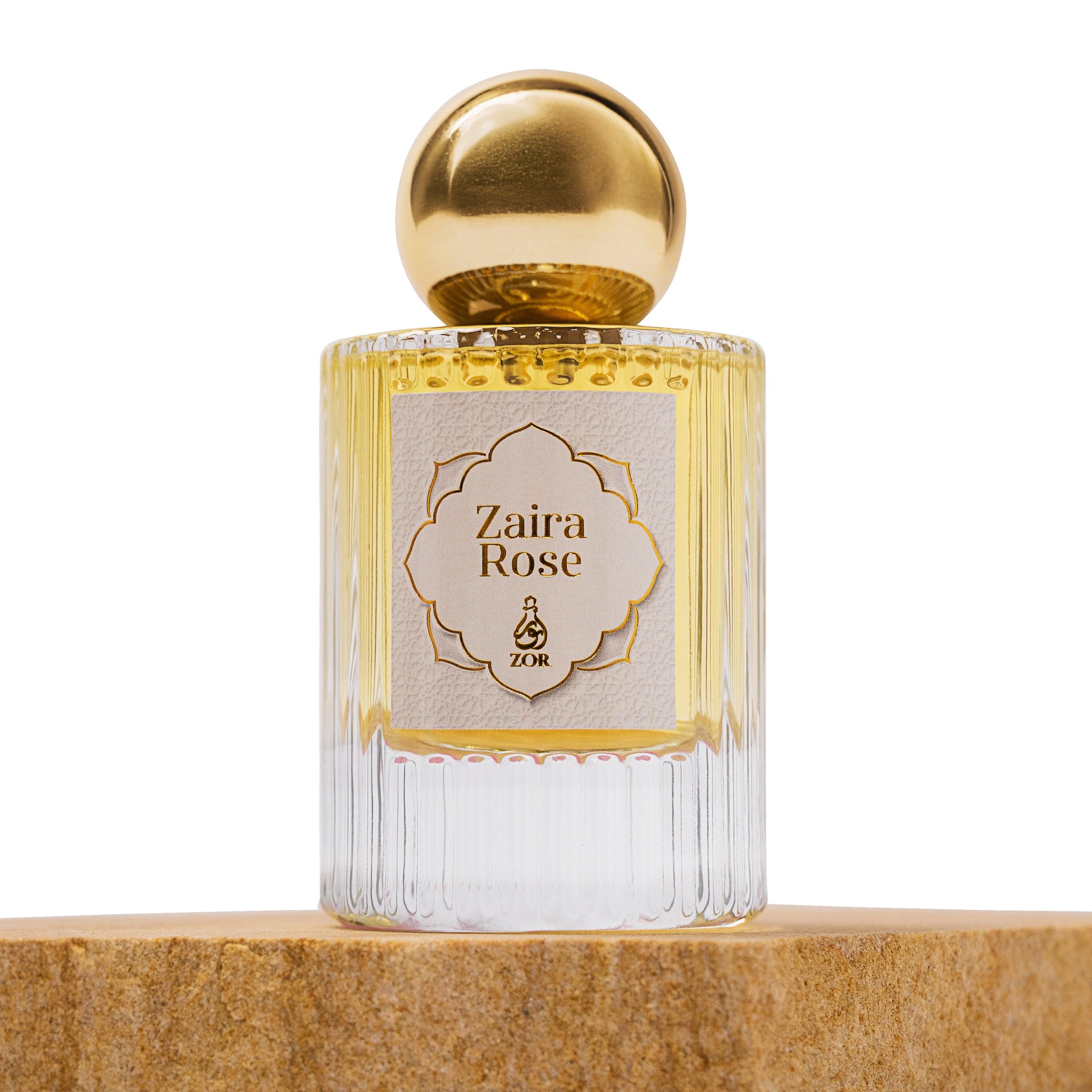 A perfume bottle labeled "Zaira Rose" by ZOR Parfums with a gold cap and a ribbed, transparent flacon sits on a wooden surface. The label is ornate with a gold border, giving the bottle an elegant appearance, hinting at notes of honey and oud within.