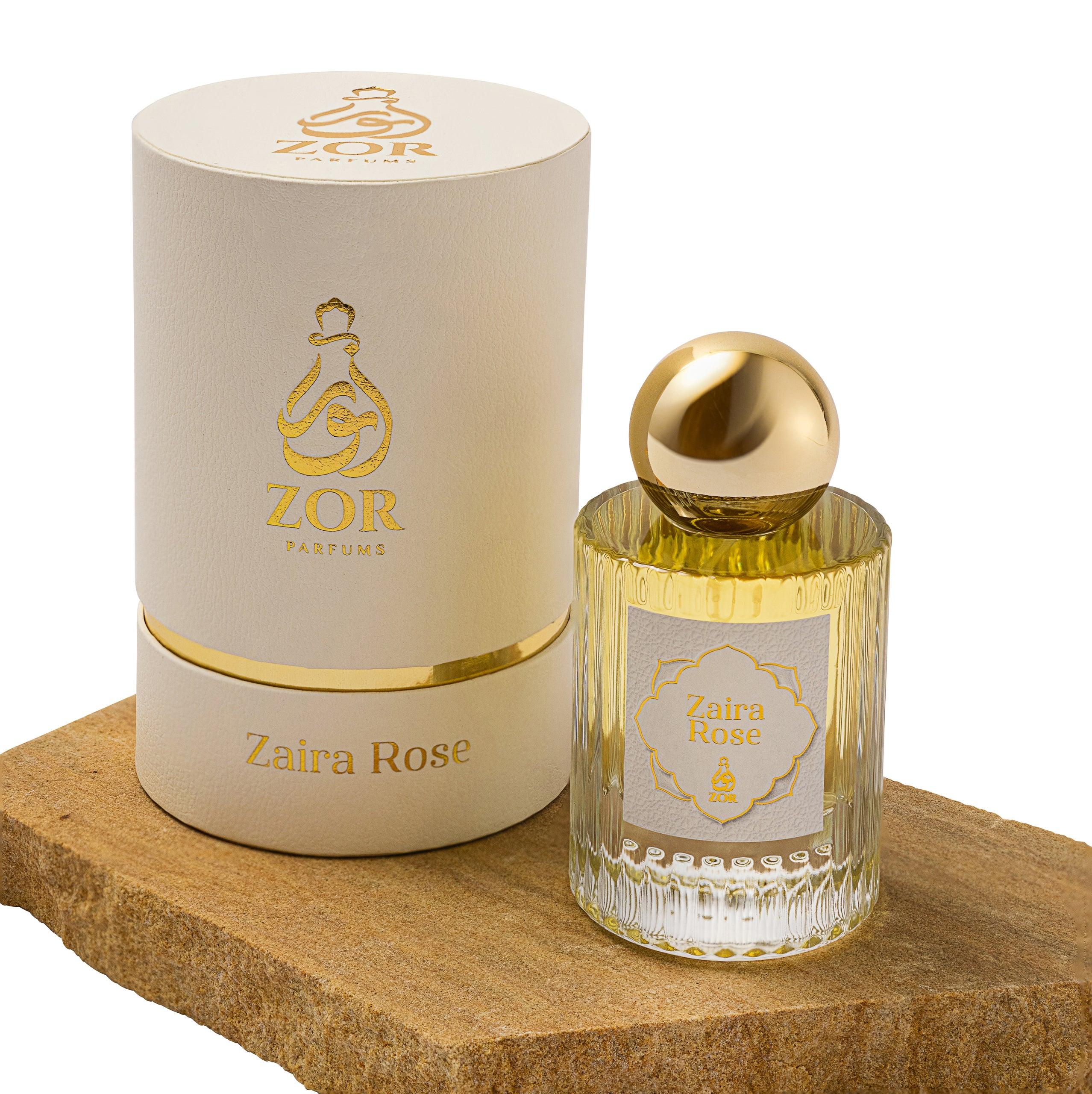 A luxury perfume bottle labeled "Zaira Rose" from ZOR Parfums is shown next to its matching cream-colored cylindrical packaging. The bottle, featuring a gold cap and elegant vertical grooves, hints at the warm, honeyed notes of oud within. Both the bottle and packaging are adorned with gold lettering and designs.