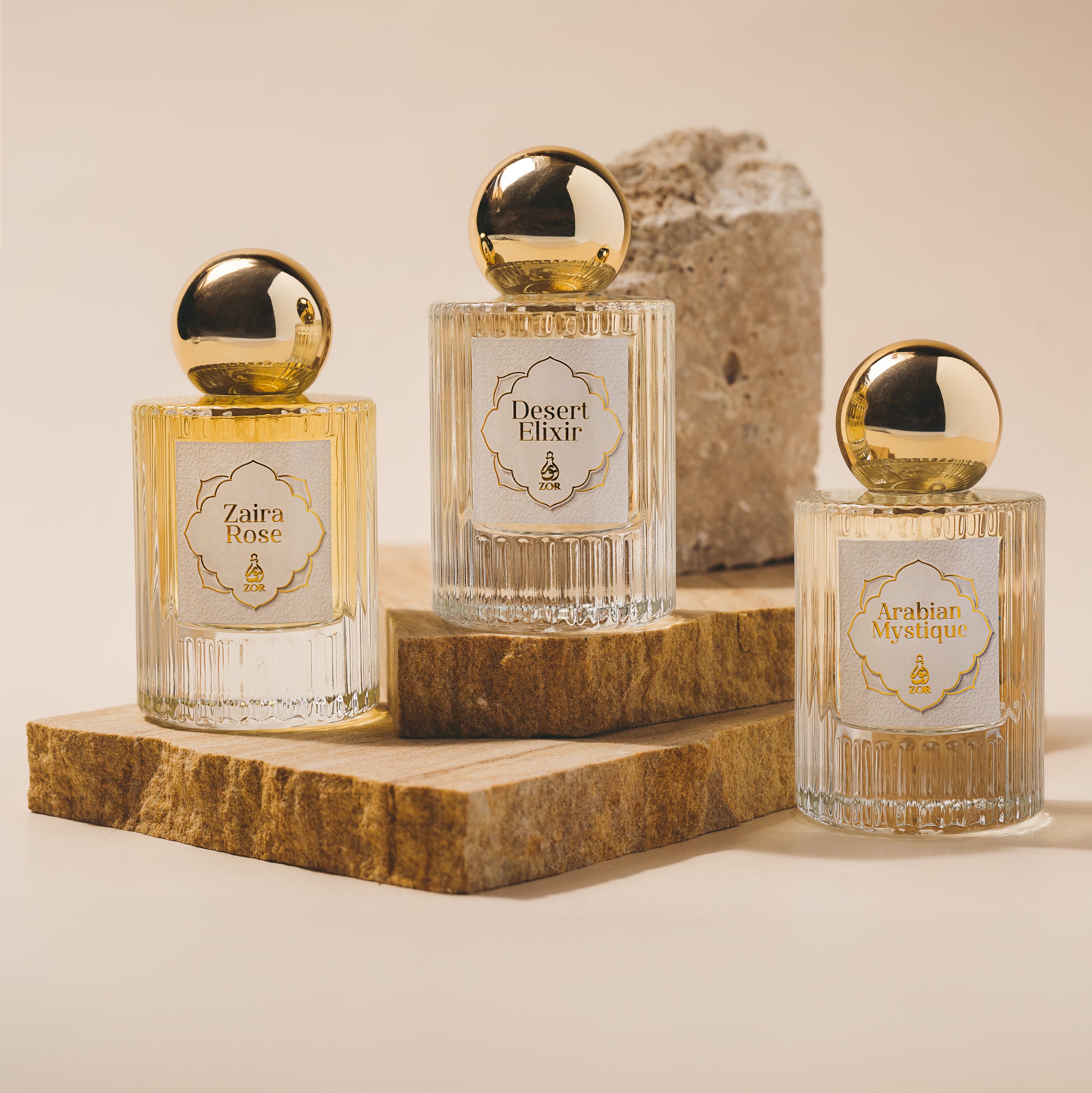 An arrangement of three captivating fragrances from ZOR Parfums, "Zaira Rose," "Desert Elixir," and "Arabian Mystique," on stone slabs. Each clear bottle boasts a gold round cap and ornate label. A textured stone block in the background enhances this elegant Discovery Set presentation by ZOR Parfums.