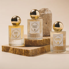 An arrangement of three captivating fragrances from ZOR Parfums, "Zaira Rose," "Desert Elixir," and "Arabian Mystique," on stone slabs. Each clear bottle boasts a gold round cap and ornate label. A textured stone block in the background enhances this elegant Discovery Set presentation by ZOR Parfums.