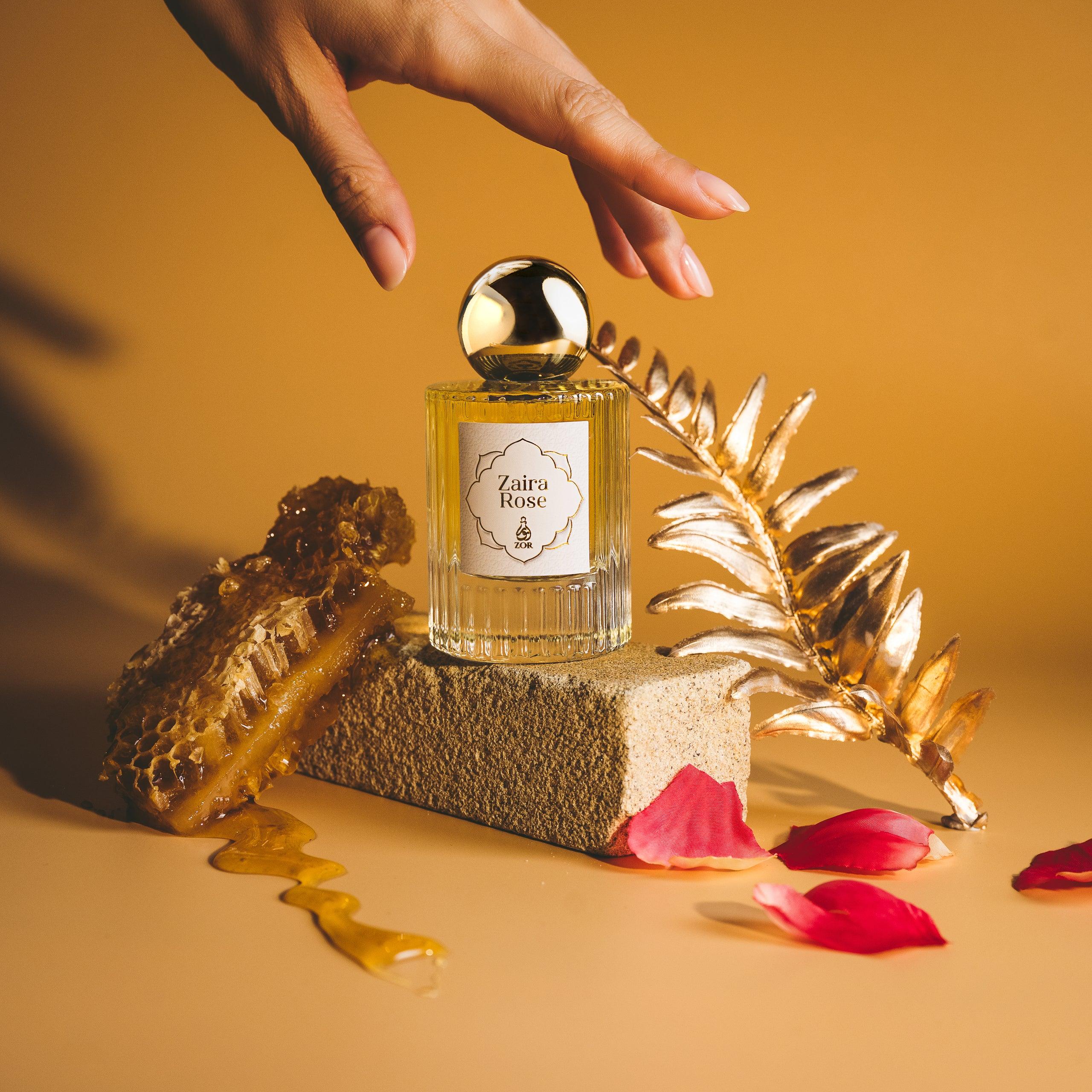 A hand reaches for a perfume bottle labeled "Zaira Rose" by ZOR Parfums adorned with a golden spherical cap. The bottle is elegantly placed on a textured block surrounded by honeycomb, gold leaf, and scattered rose petals against an orange-brown background. The scene exudes luxury with hints of oud and honey.