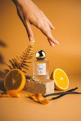 A hand reaches towards a clear perfume bottle with a gold cap, labeled "Desert Elixir." The bottle from ZOR Parfums is set on a stone block surrounded by sliced oranges, orange peels, vanilla beans, and a golden fern against a warm, beige background. Its subtle orange blossom scent captures the essence of its name.